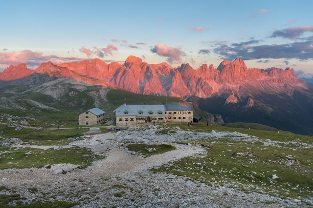 A stone mountain hut on a high meadow below peaks lit pink by sunset