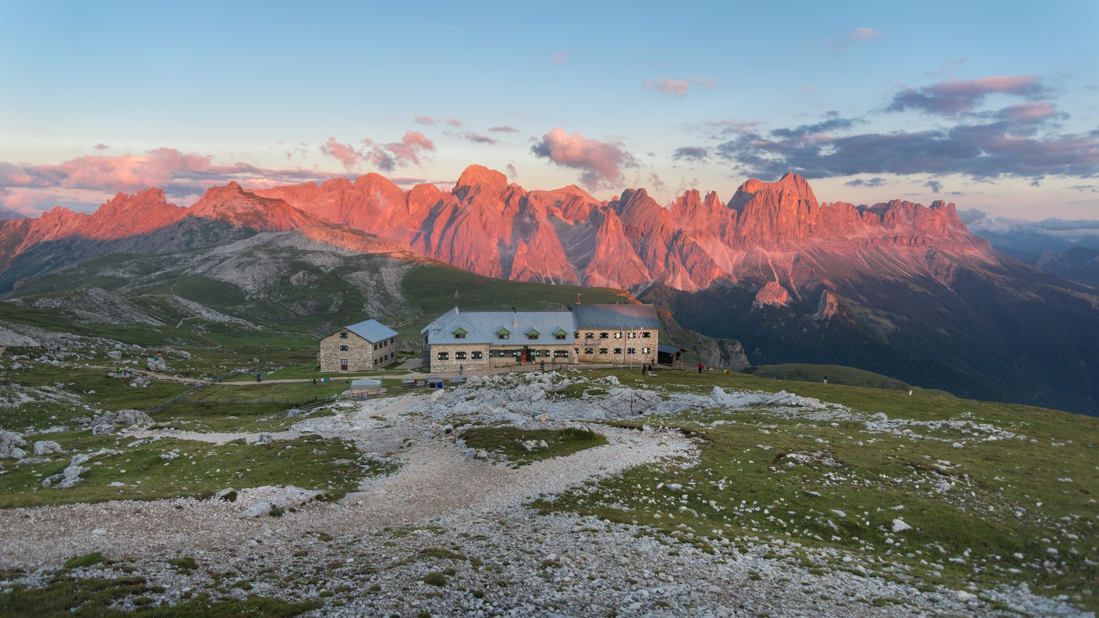 A stone mountain hut on a high meadow below peaks lit pink by sunset