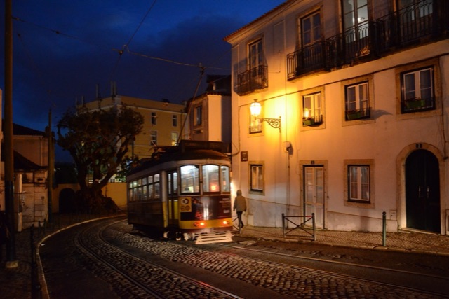 An old yellow tram rounding a cobbled bend at dusk beneath a glowing street lamp