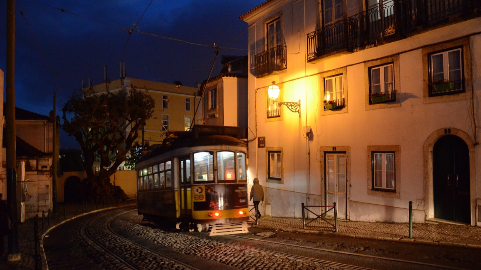 An old yellow tram rounding a cobbled bend at dusk beneath a glowing street lamp