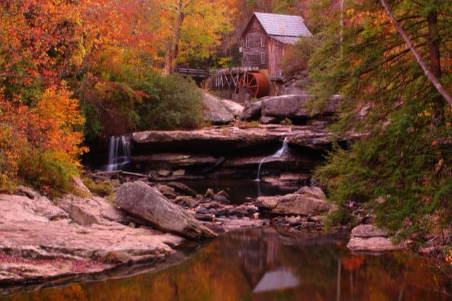 A wooden grist mill with a red water wheel above a still creek among autumn trees