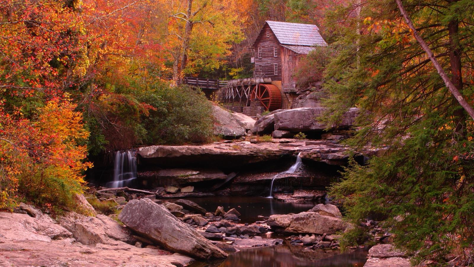 A wooden grist mill with a red water wheel above a still creek among autumn trees